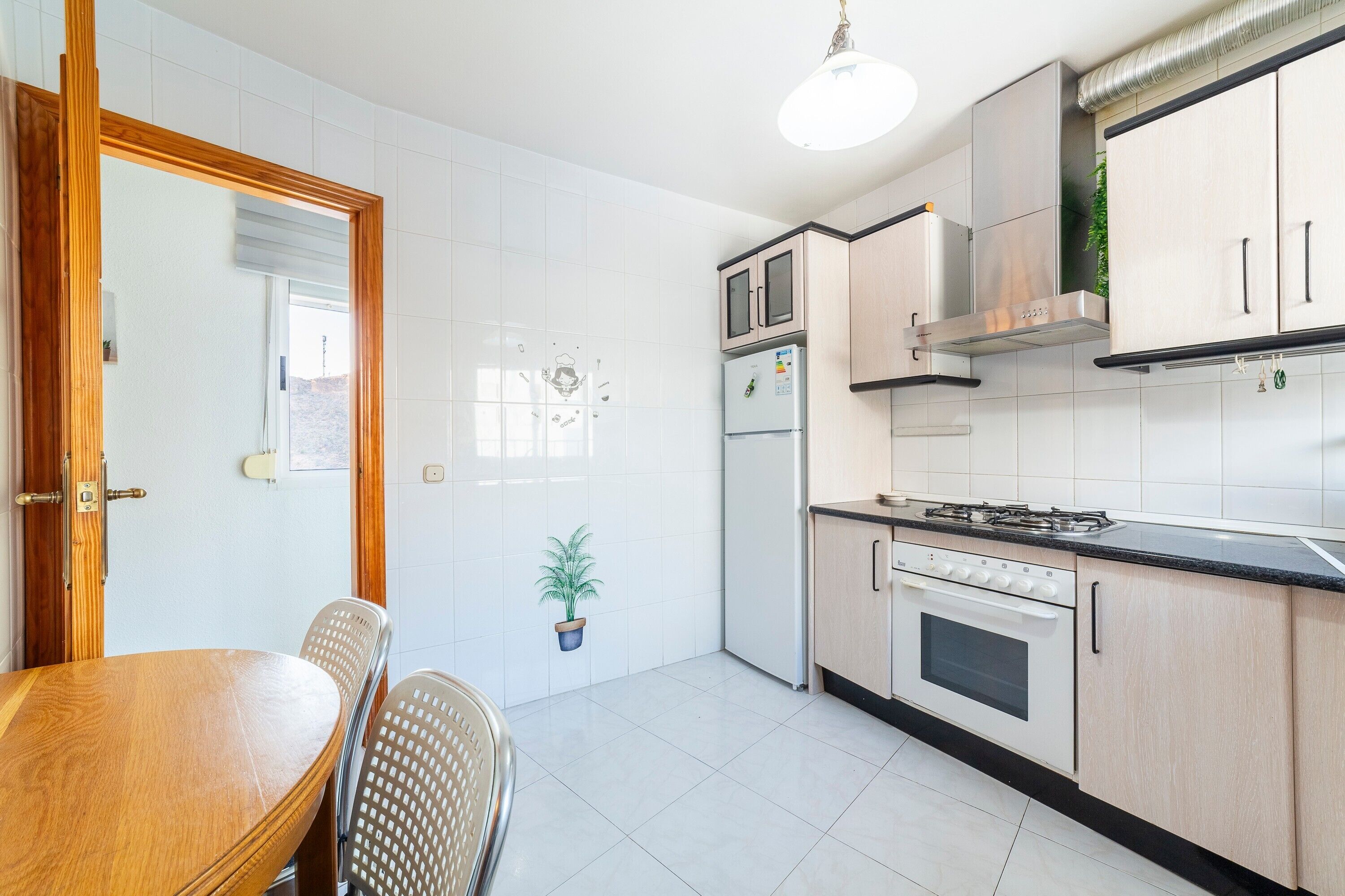 A kitchen with white tiles, a wooden dining table, chairs, a stove, and cabinets.