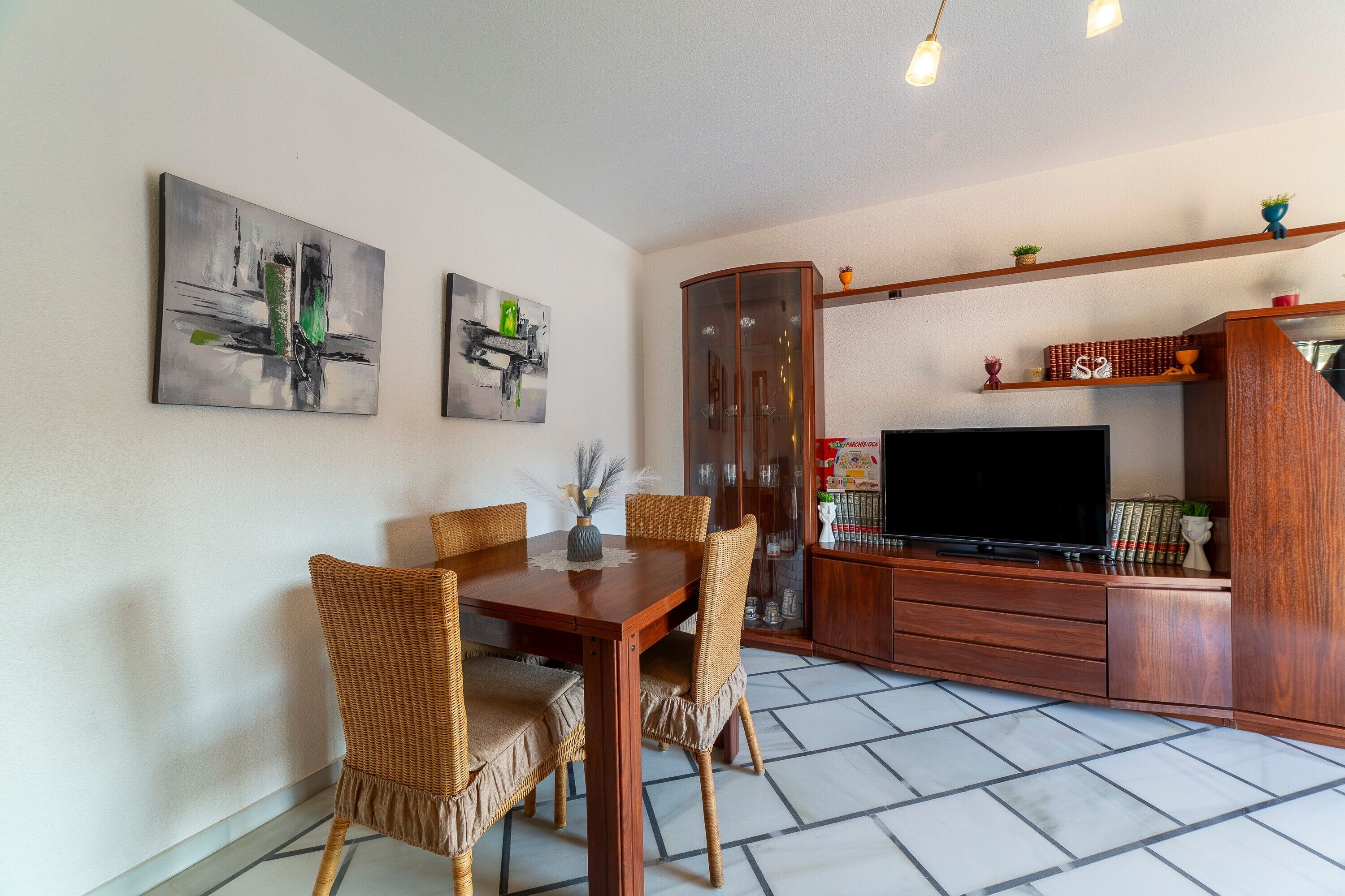 Un comedor con una mesa de madera y sillas de mimbre, un mueble con televisor y obras de arte en las paredes.