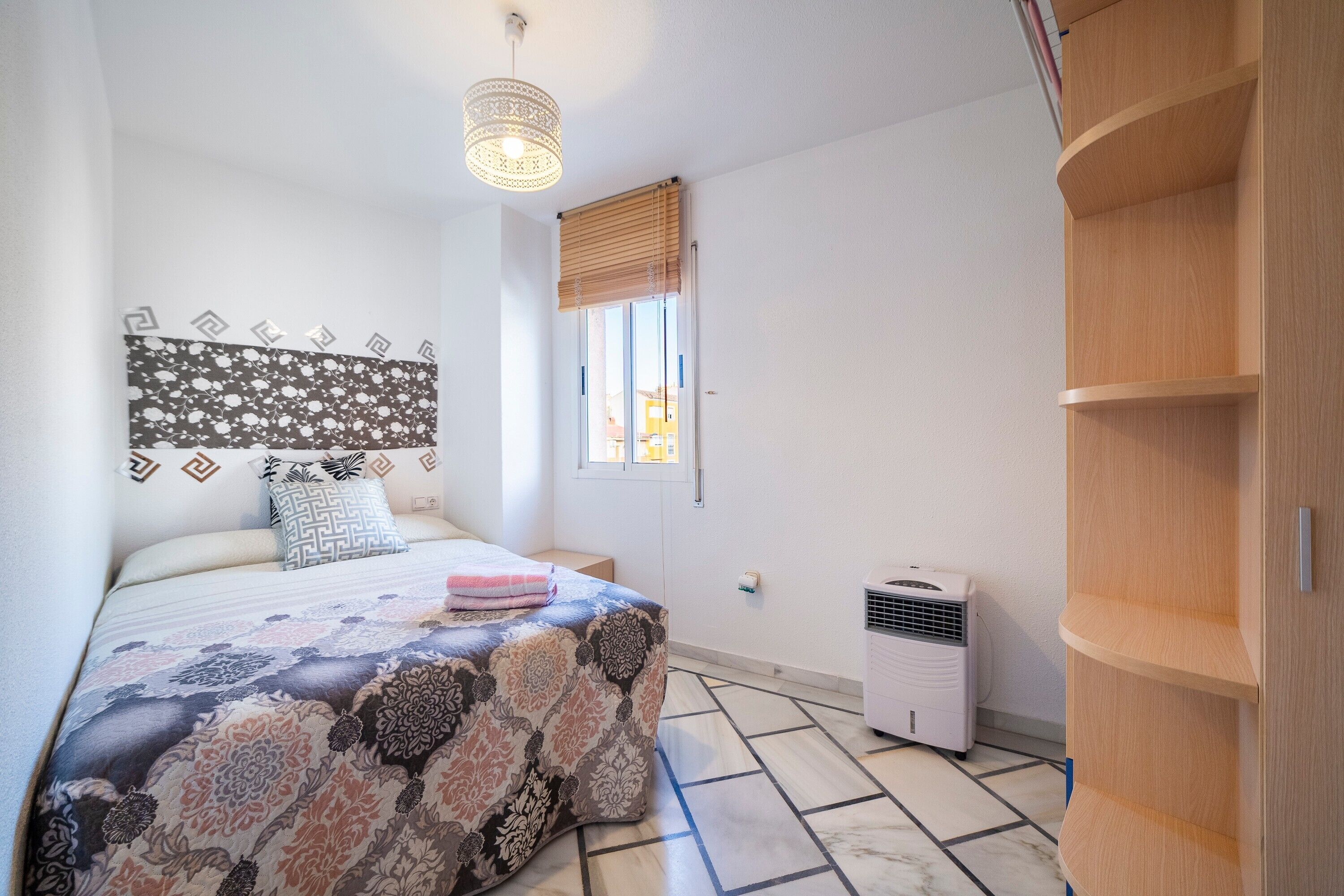 Un dormitorio con una colcha estampada, un cabecero con diseño geométrico, una lámpara de noche, una ventana con persianas y un equipo de aire acondicionado instalado.