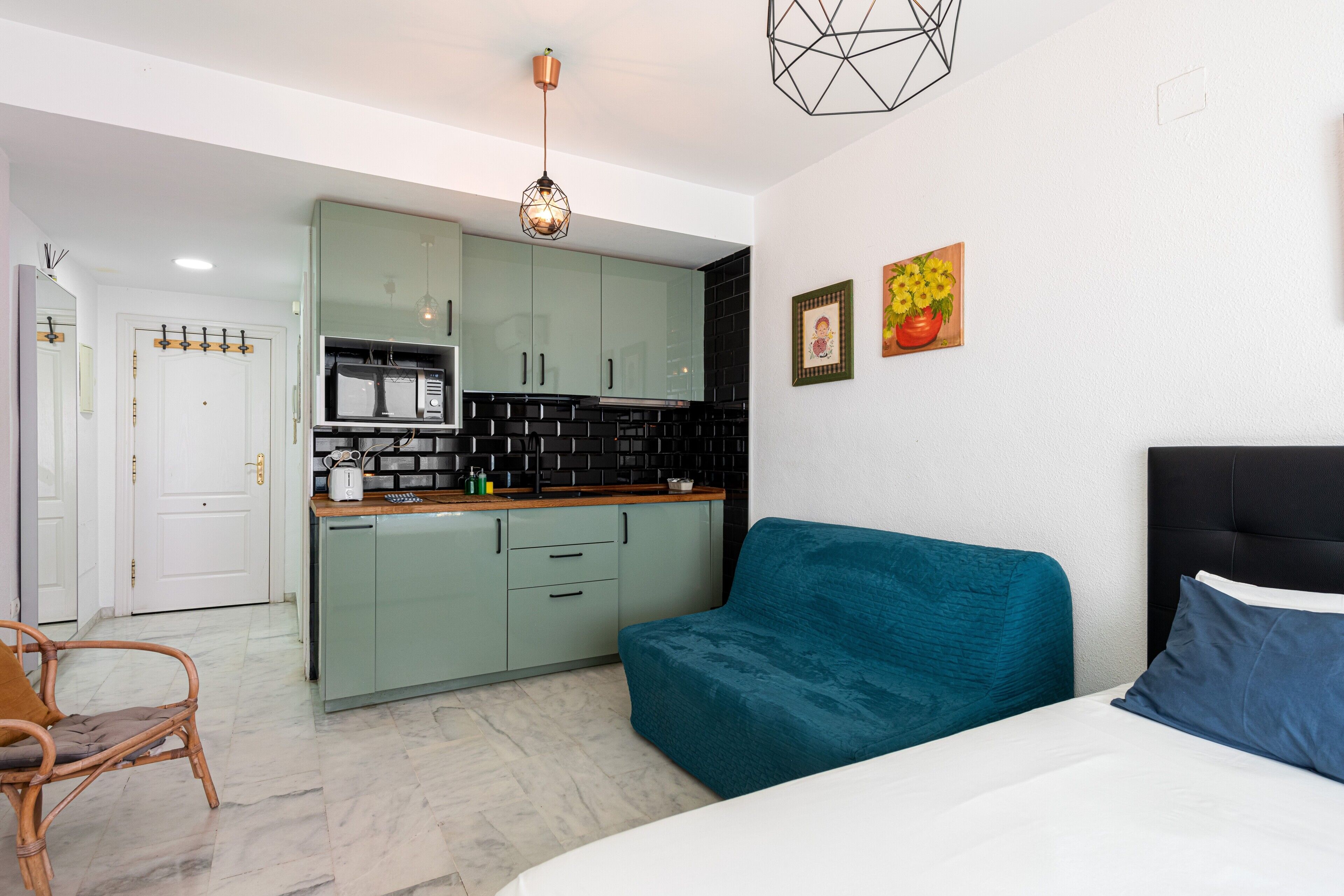 A compact room with a kitchenette, a teal sofa, a bed with blue pillows, and a hanging light fixture.