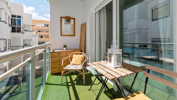 A balcony with a wooden table, a wicker chair, a white lantern, and a mirror on the wall.