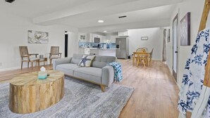 Een ruime woonkamer met een grijze bank, houten stoelen, een ronde salontafel en een keuken op de achtergrond.