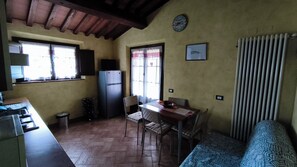A compact kitchen with a refrigerator, a dining table with chairs, a window with curtains, and a radiator.