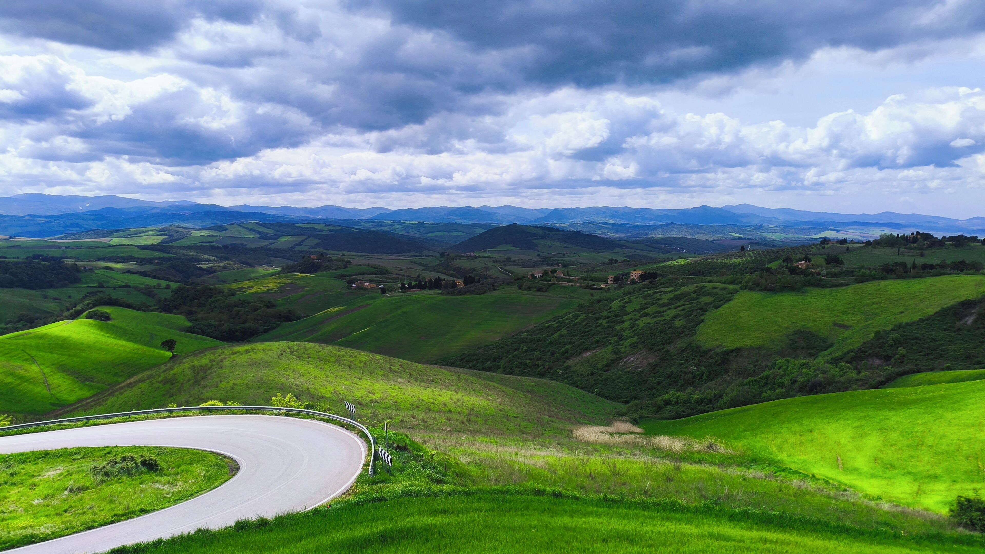 A winding road through rolling green hills with distant mountains under a cloudy sky.