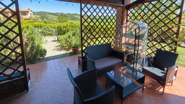 A covered patio with wicker furniture, a glass table, and a view of a garden and hills.