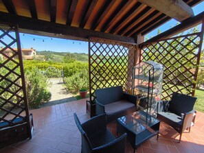 A covered patio with wicker furniture, a glass table, and a view of a garden and hills.