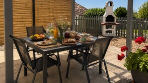 Un espace extérieur abrité, aménagé pour prendre les repas, avec une table dressée pour le petit-déjeuner, comprenant des fruits, du pain et des boissons.