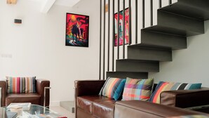 A modern living room with a brown leather sofa, colorful striped pillows, a glass coffee table, and a staircase with black railings. - Lodge du Hautacam (Ayros-Arbouix)