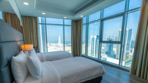 A modern hotel room with a large bed, a headboard, a lamp, and a view of the cityscape through large windows. - S Plaza Suites Hotel (Manama)
