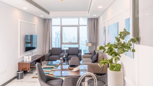 A modern living room with a large window, a flat-screen TV, grey sofas, a coffee table, and a potted plant. - S Plaza Suites Hotel (Manama)