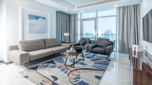 A modern living room with a large sofa, a smaller sofa, and a coffee table. There is a large window with curtains, a painting on the wall, and a view of the city. - S Plaza Suites Hotel (Manama)