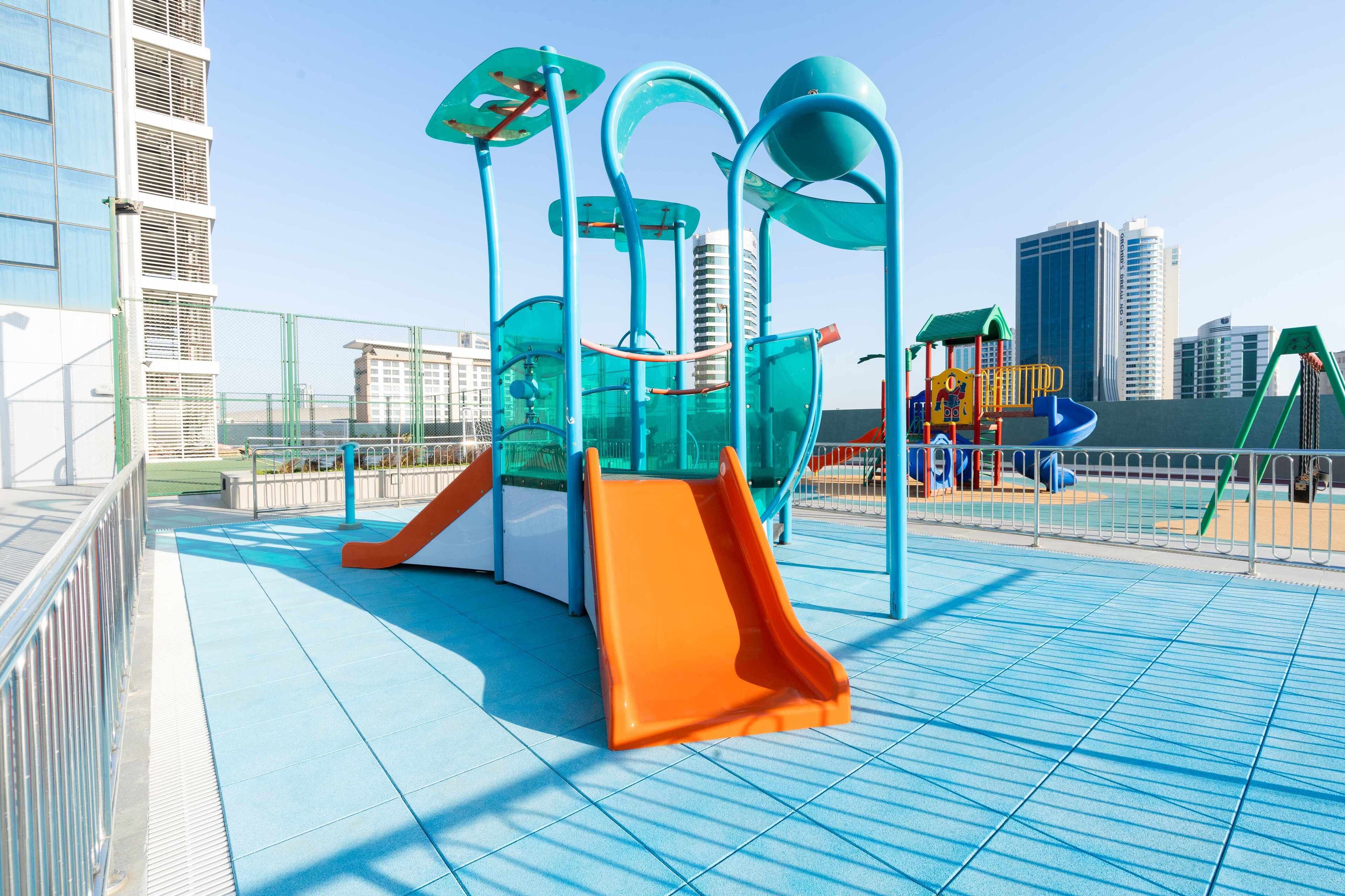 Un patio de juegos con toboganes y estructuras para escalar, rodeado por una cerca y edificios al fondo.