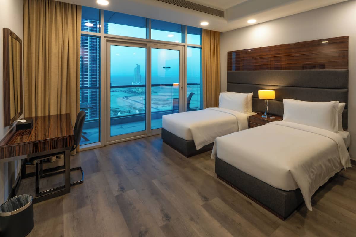 A hotel room with a large window, two beds, a desk, and a chair.