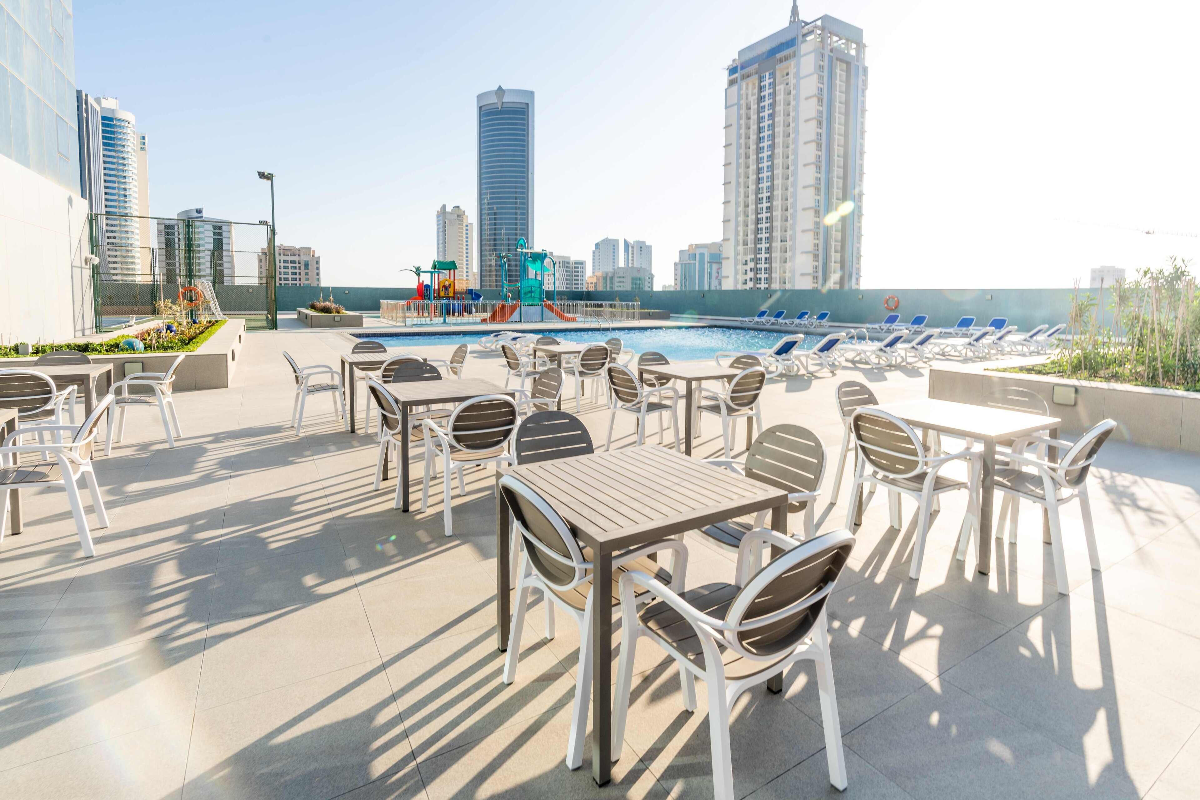 A rooftop pool area with lounge chairs and tables, surrounded by high-rise buildings.