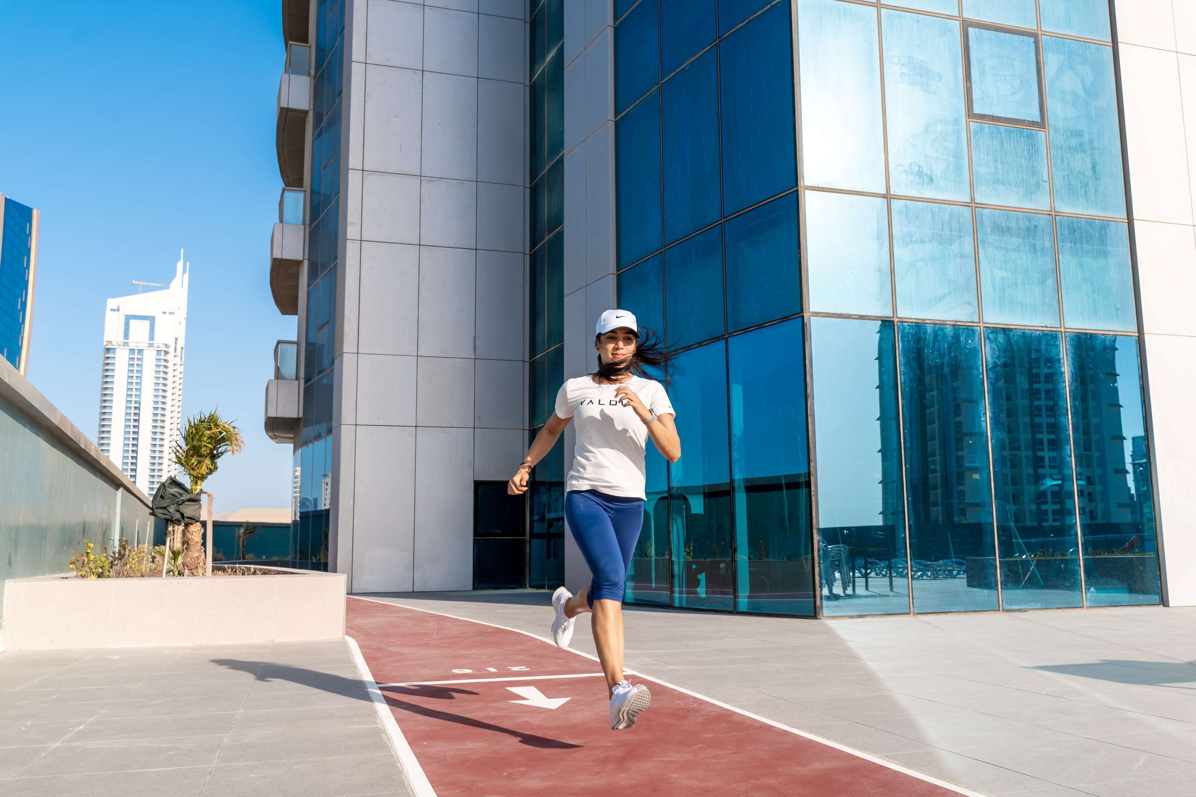 A person running on a red track in front of a modern building with large glass windows.