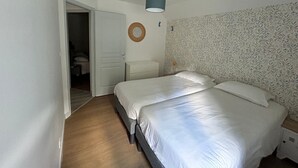 Quarto com duas camas, piso de madeira, porta branca, luminária fixada na parede e papel de parede estampado.