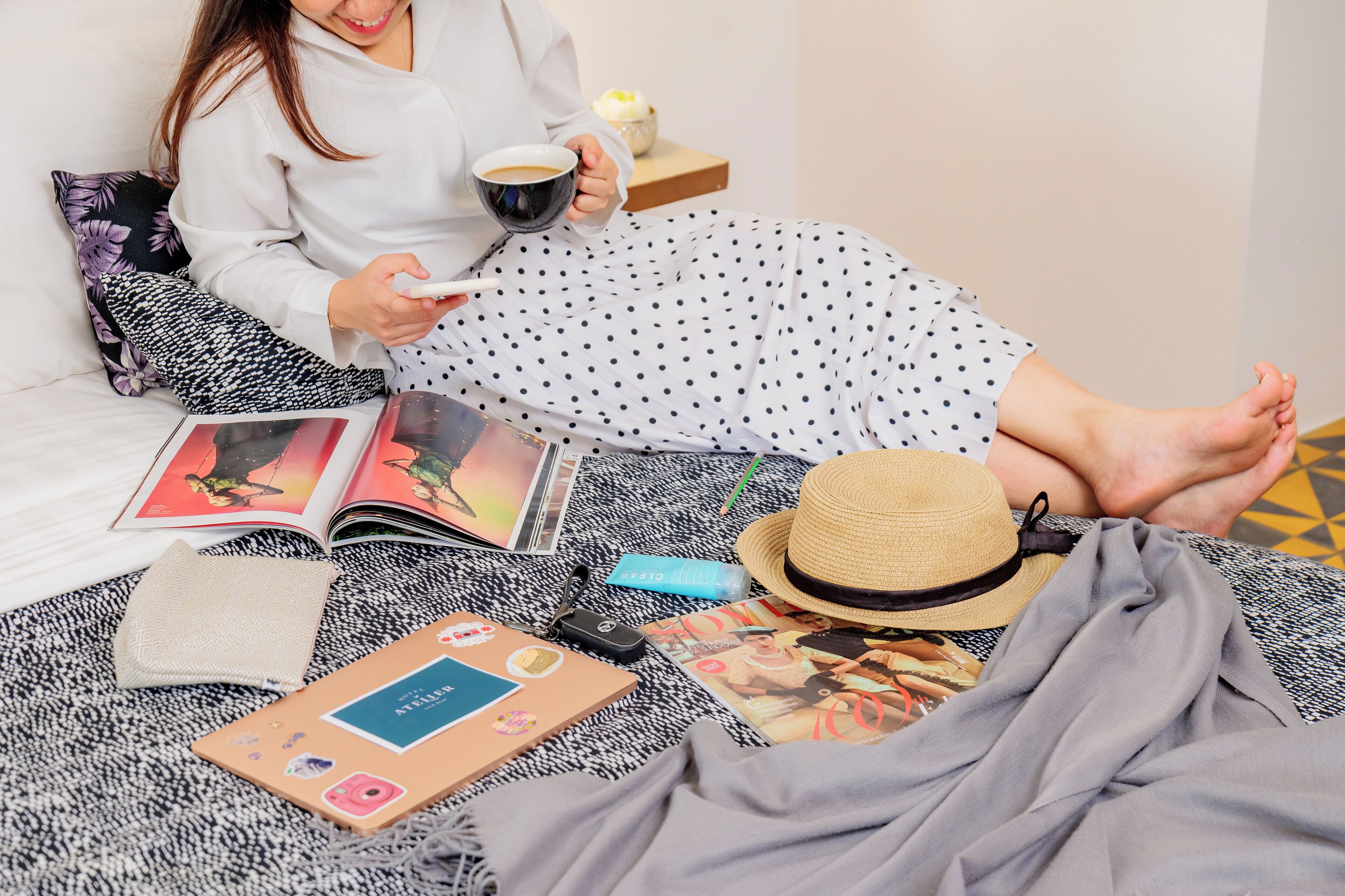 Una persona sentada en una cama con un libro, una taza y un sombrero.