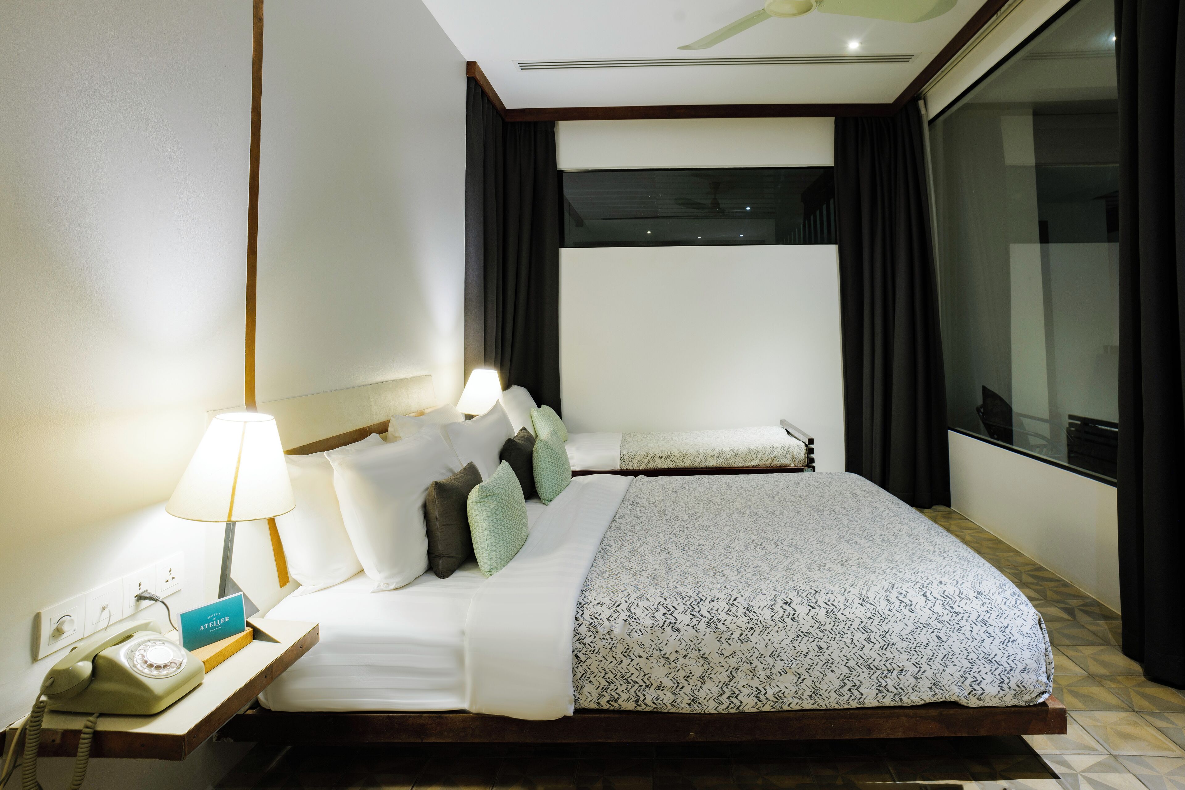A hotel room with a bed, a nightstand with a lamp, a telephone, and a view of the outside through a large window.