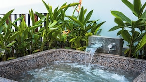 A rectangular hot tub with a speckled stone exterior, surrounded by lush green foliage. - Atelier Hotel (Siem Reap)