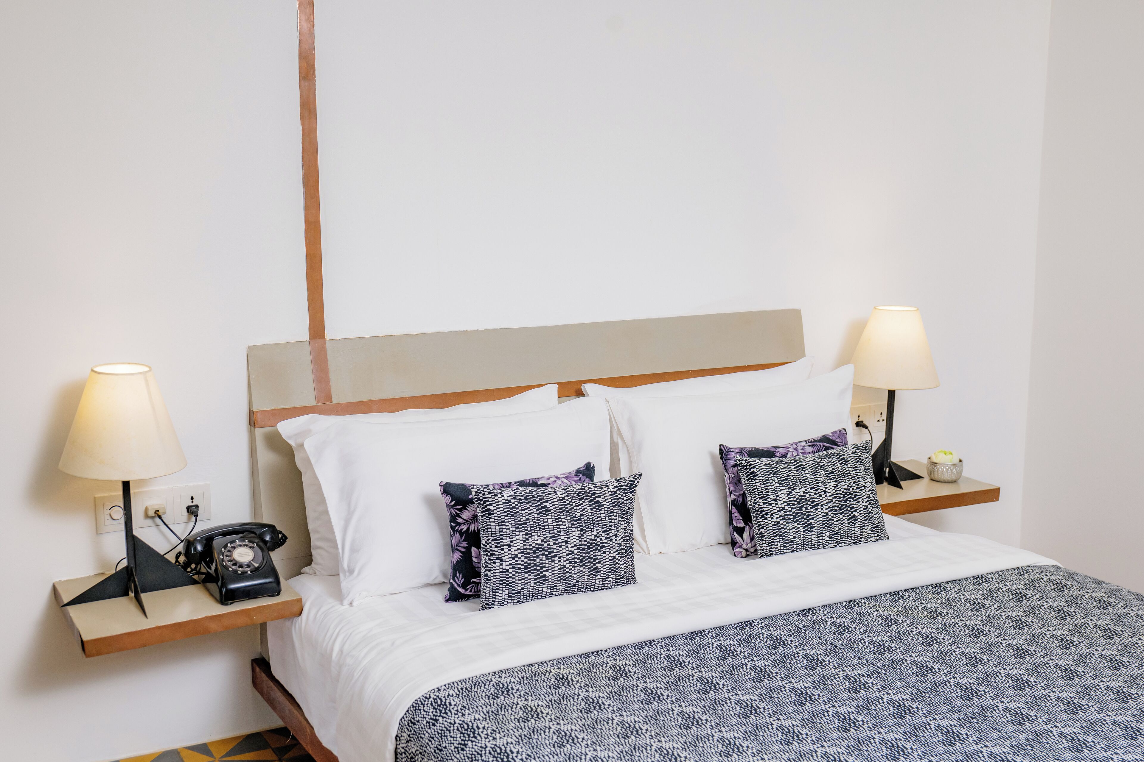 A neatly made bed with a patterned bedspread, two bedside lamps, a telephone, and a small table with a lamp and a decorative item.
