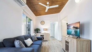 A modern living room with a sofa, TV, wooden cabinet, ceiling fan, and a clock on the wall. - Seaside Serenity—Family Escape with Private Spa (Machans Beach)
