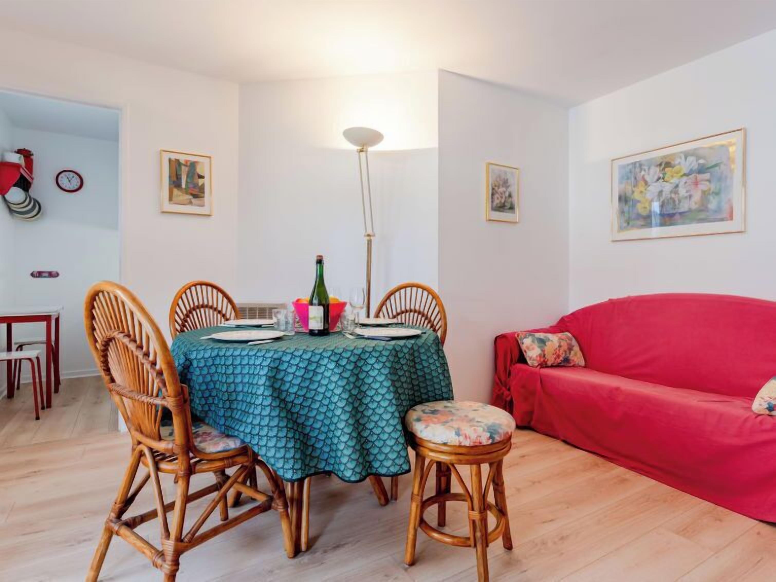 Un comedor con una mesa redonda cubierta con un mantel verde, rodeada de sillas de mimbre. Se observa un sofá rojo al fondo y una lámpara de pie sobre la mesa.