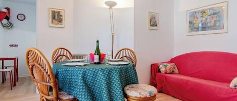 Un comedor con una mesa redonda cubierta con un mantel verde, rodeada de sillas de mimbre. Se observa un sofá rojo al fondo y una lámpara de pie sobre la mesa.