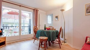 A dining area with a table set for two, wooden chairs, a green tablecloth, and a view through a sliding glass door to an outdoor balcony with a white fence and a pink umbrella. - Le Clos Mathilde by Interhome (Cabourg)