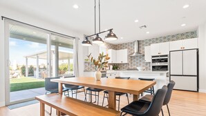 A modern kitchen with white cabinets, a central island, and a dining area with a wooden table and chairs. - Rockslide House - New build, resort style pool, endless amenities (3 miles to festivals) (Coachella)
