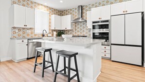 A modern kitchen with white cabinets, a central island with stools, a built-in oven, and a stainless steel range hood. - Rockslide House - New build, resort style pool, endless amenities (3 miles to festivals) (Coachella)