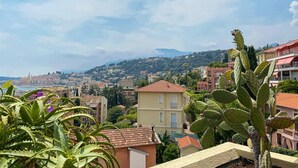 Une ville côtière à l’architecture méditerranéenne, avec des bâtiments aux toits de tuiles rouges et un grand cactus bien visible au premier plan.