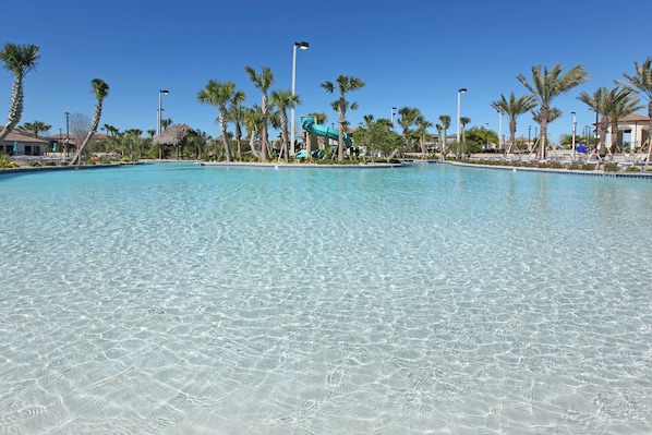 A clear swimming pool with a water slide, surrounded by palm trees and a thatched-roof structure.
