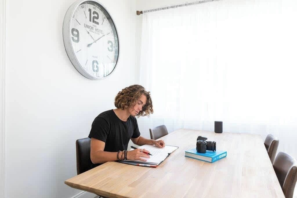 Una persona sentada a una mesa de madera en una habitación con un gran reloj de pared, una ventana con cortinas, y un libro azul y una cámara sobre la mesa.