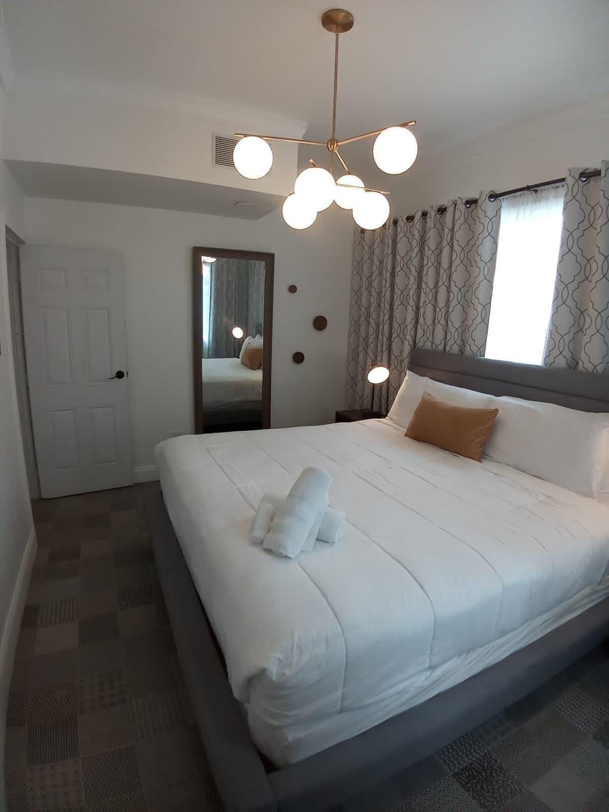 A neatly made bed with a white comforter and a brown pillow, a modern chandelier, and a large mirror on the wall.