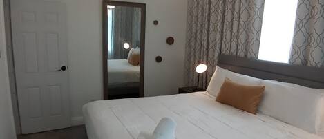 A neatly made bed with a white comforter and a brown pillow, a modern chandelier, and a large mirror on the wall.