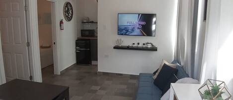 Un salon moderne avec un téléviseur à écran plat, un canapé, une table à manger et un coin cuisine à l’arrière-plan.