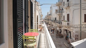 A narrow street with buildings on either side, a balcony with a table and chairs, and a shop with items displayed outside.