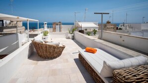 Eine Dachterrasse mit einem Korbwicker-Korb, einer weißen Sitzbank mit Kissen und Blick aufs Meer.