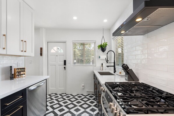 A compact kitchen with white cabinetry, a black and white patterned floor, a gas stove, and a window with blinds.