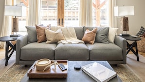 A modern living room with a grey sofa, multiple cushions, a coffee table with a tray and remote control, and a book titled "American." - Town Center | Hot Tub | Ski Shuttle (Big Sky)