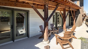 A covered outdoor seating area with wooden chairs and a table, a wooden pergola structure, and a sliding glass door leading to a patio. - Town Center | Hot Tub | Ski Shuttle (Big Sky)