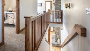 A multi-level interior with a wooden staircase, a stone accent wall, and a view into another room with a bed and a lamp. - Town Center | Hot Tub | Ski Shuttle (Big Sky)