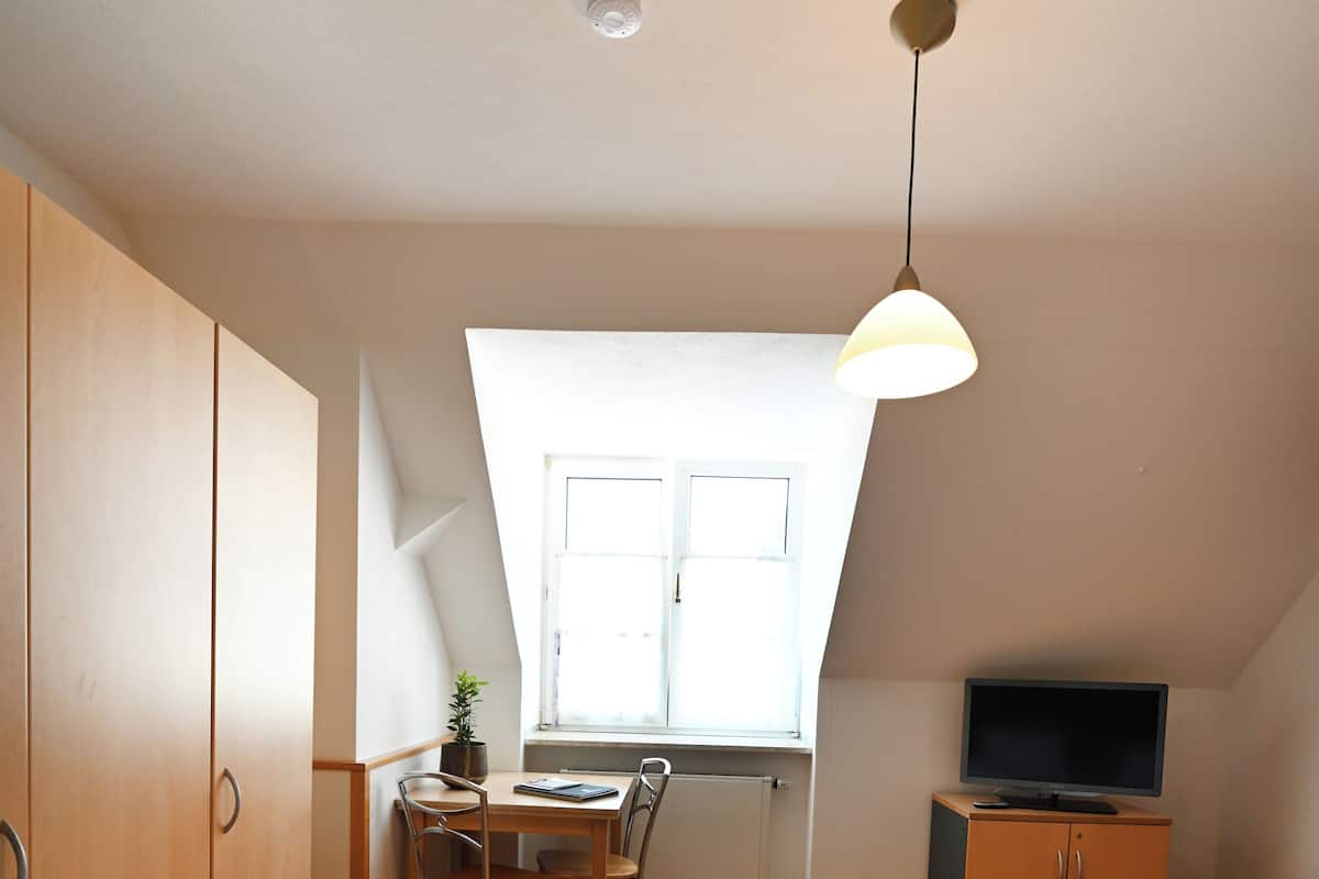 Una habitación con una cama, un escritorio con silla, un televisor sobre un mueble bajo y una ventana que deja entrar luz natural.