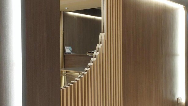 A modern hallway with a wooden floor, a curved mirror, and vertical wooden slats. - Hotel Grand Galaxy (Yangon)