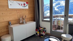 A modern room with a wooden wall, a white cabinet, a colorful cube, a fluffy stool, a round table with a remote, and a window with a snowy mountain view.