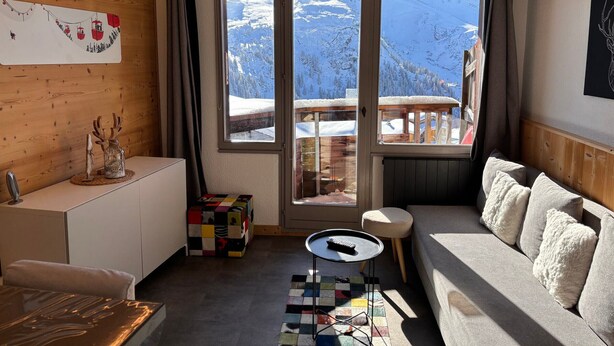 A cozy room with a wooden interior, a sofa, a small table, and a view of snowy mountains through large windows.