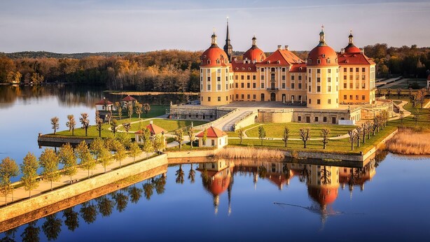 Ein großes, historisches Gebäude mit mehreren Kuppeln und einem zentralen Turm, umgeben von einem ruhigen See und einem gepflegten Garten.