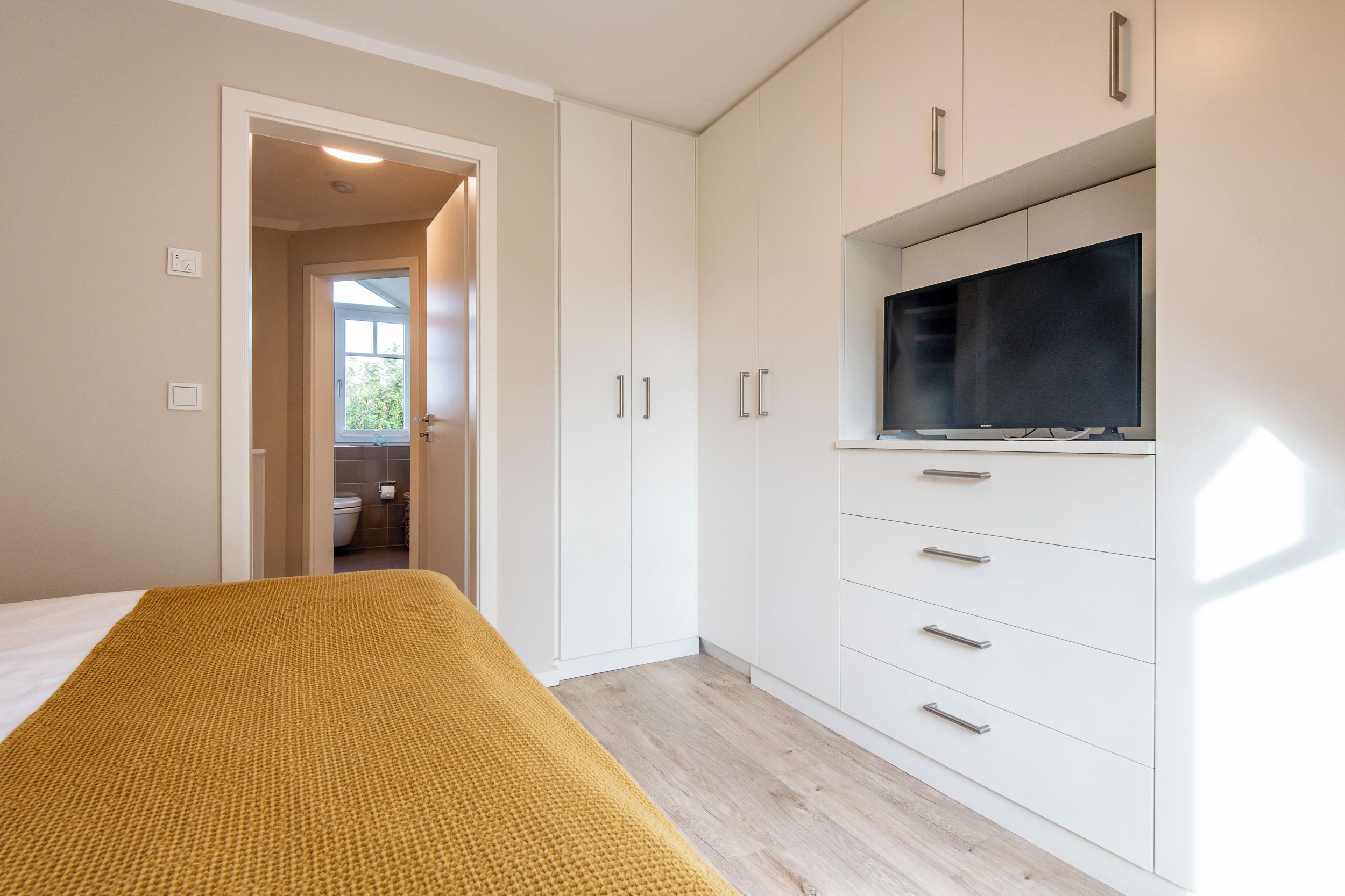 Quarto com cama, guarda-roupa, televisão e banheiro visível por uma porta aberta.