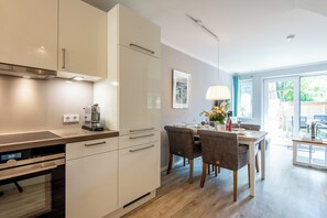 A modern kitchen with white cabinets, a built-in oven, and a dining area with a table and chairs. - Zentral. Familie. Neubau. Objekt ID. Dbs/12d (Westerland)