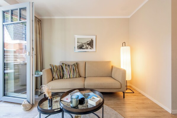 A living room with a sofa, a coffee table, a lamp, and a framed picture on the wall. - Zentral. Familie. Neubau. Objekt ID. Dbs/12d (Westerland)